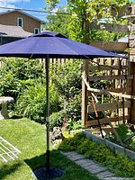 Full view of navy blue Corona branded patio umbrella with metal pole and wrought iron umbrella stand on green lawn with garden background.