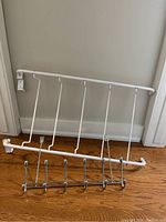White metal over-the-door clothes hanger with five vertical hooks and chrome metal over-the-door clothes hanger with five hooks, both placed on the floor against a wall.