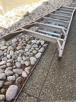 Photo of aluminum extension ladder lying on a paved surface next to rocks, showing side and bottom supports and rungs.