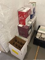 Corner view capturing multiple cardboard boxes filled with empty wine bottles, stacked against a white wall with visible texture.