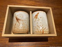 Two Japanese teacups in cream and rust tones placed inside a wooden kiribako box with a divider.
