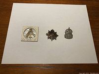 Front view of three cap badges on a white sheet: Royal Canadian Army Pay Corps badge in gold tone, Canadian Rifles of Canada badge in brass tone, and WW2 ARP badge in silver tone.