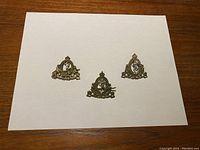 Three gold-toned Royal Canadian Corps of Signals cap badges displayed on white paper, showing front side.
