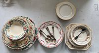 Overview of antique plates and silverware set including multiple ceramic plates of various sizes and floral patterns, and metal spoons.