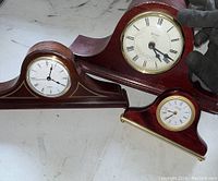 Three vintage mantel clocks arranged on a surface showing tops and faces with wood finishes and Roman numerals, providing overall lot view.
