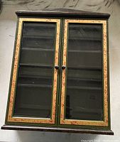 Front view of curio cabinet with closed glass doors showing black and gold-red trim painted wood.