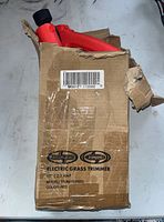 View of the electric grass trimmer partially visible inside a worn cardboard box, with box label showing brand and model information.