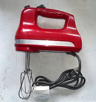 Front and side view of red KitchenAid 3 Speed Hand Mixer with two metal beaters and cord wrapped beside it on a white surface.