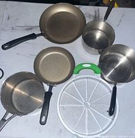 Top-down image showing five metal pans including frying pans and saucepans with black and green handles on a white surface.