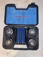 Open black hard plastic carrying case with blue insert labeled 'PROSport'. Inside are two adjustable dumbbell bars with blue foam grips and four metal weight plates.