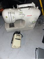 Side angle view of white Singer Inspiration sewing machine with foot pedal on grey surface. Shows the main body, stitch selector dial, and foot pedal details.