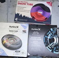Three boxed inflatable snow tubes: Crescent Moon 60-inch tube in red and purple, Hurley 48-inch tube with colorful design, and Hurley black and blue tube.