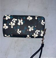 Exterior photo showing black wallet with white floral print and zip-around zipper with attached wrist strap.