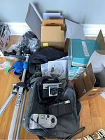 Overview of all cameras and equipment arranged together showing the black Kodak Instamatic X-15, Lumix camera, Sony Cyber-shot, Nikon lens box, tripod and manuals