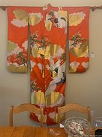 Full front view of a bright orange kimono hanging on a wooden rod, showing large crane and pine tree embroidery with gold cloud accents.