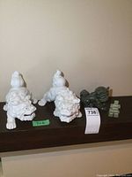 Pair of white porcelain Foo dog statues next to smaller alabaster lion and green inukshuk figurines placed on a wood shelf.