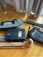 Photo showing Lohanu ukulele case, black hard clarinet case, two Yamaha branded beige recorder cases, white metronome, and guitar stands all on wooden floor.