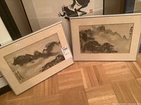 Photo showing two framed Asian watercolour paintings side by side, laid on wooden floor: mountain and tree landscapes, beige mattes, light-colored frames.
