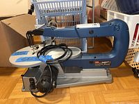 Side view of Ryobi SC162 VS scroll saw showing the entire tool, with blue arm, metal base, power cord coiled on the base, and a piece of blue tape labeled 'SELL'.