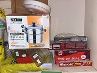 White serving trays stacked behind boxed stainless steel pot and several rolls of kitchen wraps sample including plastic wrap, parchment paper, and foil.