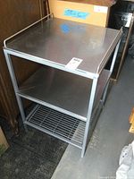 Front and side view of the stainless steel workstation showing top surface, middle shelf, bottom wire shelf, and wheels on one end.