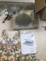 Full view of Dual CS 514 turntable with clear dust cover closed, power cord beside it and manual in plastic wrap in front.