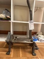 Side angled view of the vintage French fry cutter showing the handle, blade assembly and four supporting legs on wooden floor with white storage cubbies background.