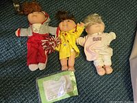 Three Cabbage Patch Kids dolls lying on carpet, dressed in different outfits, with certificate included