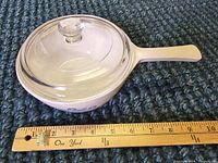 White ceramic CorningWare saucepan with long handle and blue cornflower pattern alongside a wooden ruler for size reference.