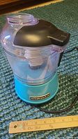 Side view of KitchenAid chopper showing turquoise base with KitchenAid logo, clear container, and lid. Ruler present for scale indicating about 7 inches tall.