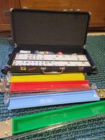 Photo showing open black carrying case with Mahjong tiles arranged inside, colorful chips, dice, and four colored plastic racks outside case.