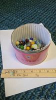 Photo showing the mixed glass marbles inside the pink Almond Roca tin with a ruler for size reference.