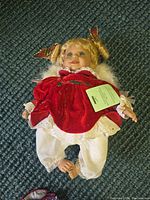 Full view of the doll lying on carpet with blond hair, red velvet dress, white bloomers, and feathered wings, showing overall condition and attire.