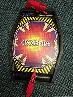 Top-down view of the Crossfire game board showing the playing surface with the game name and the two red plastic shooters at each end. Metal balls are visible near one red puck.