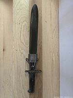 Full view of the WW2 US bayonet showing black handle and metal blade with scabbard beneath it on wooden floor.