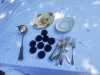 A collection of items laid out on white fabric including ten amber glass floor protectors, a silver plate soup ladle, a floral decorative plate, a white leaf-patterned plate, silver-tone forks, and vintage wooden clothespins.