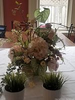 Large artificial floral centerpiece arrangement with pink roses, cream hydrangeas, white blossoms, large green heart-shaped leaves and berry branches in a cream decorative pot, along with small potted green grass plants.