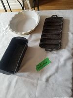 Cream-colored ceramic pie plate, cast iron cornbread mold, and metal bread pan on a white tablecloth.
