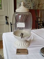 Full view of the large glass beverage dispenser on a table, showing ceramic base, lid, and label
