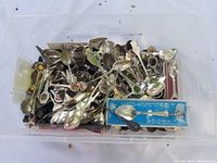 Plastic container filled with a large quantity of collectible spoons, showing a variety of designs, colors, and metal finishes.