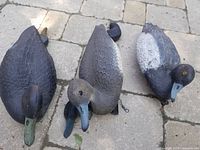 Three full decoy ducks lying on stone pavement. Ducks vary in color from black to grey with some yellow eye detail.