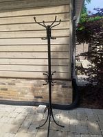 Black metal coat rack standing outdoors on a stone patio, showing its full height and multiple hooks.