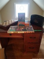 Photo shows Travel Scrabble case in black, Pictureka box, Chess set with wooden board, Clue box, and Charades box displayed on wooden desk under window light.