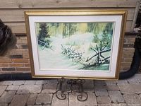 Original oil painting of a snowy forest with a wooden fence, in a gold frame on a brick patio.