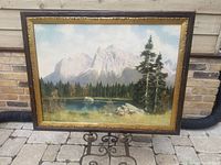 Full view of framed oil painting outdoors on stone patio, showing mountain, lake, trees in the landscape.
