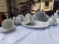 Photo showing full set of white porcelain Royal Doulton Cadence dishes including cups, saucers, plates and platter arranged on a white cloth outdoors, with a tree in the background.