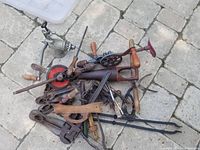 A pile of vintage hand tools lying on paving stones. Visible items include hand drills, braces, saw frame, clamps and other metal and wooden hand tools.