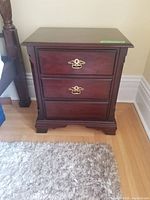 Front view of a dark wood bedside table showing three drawers, two brass handles visible, some wear seen on drawer surfaces.