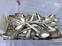Large plastic bin filled with numerous silver-tone souvenir spoons piled together, visible intricate detail on handles with state and Presidential motifs.