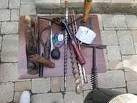 Top view of assorted hand tools on a wooden board, including a wooden-handled plane, wooden-handled spiral auger drill bits, a manual hand drill handle, chisel, a spade bit, and other metal tools.
